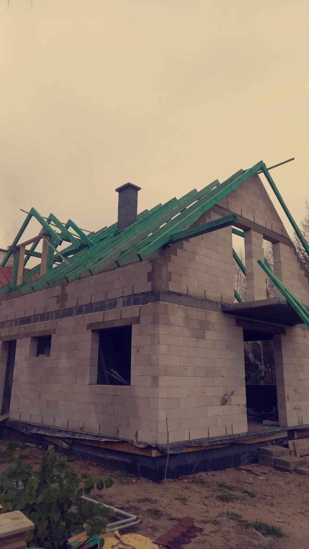 Wznoszenie dwukondygnacyjnego domu z białych bloczków, widoczne ściany, otwory okienne i drzwiowe oraz konstrukcja dachu z zielonych łat, komin w stanie surowym.