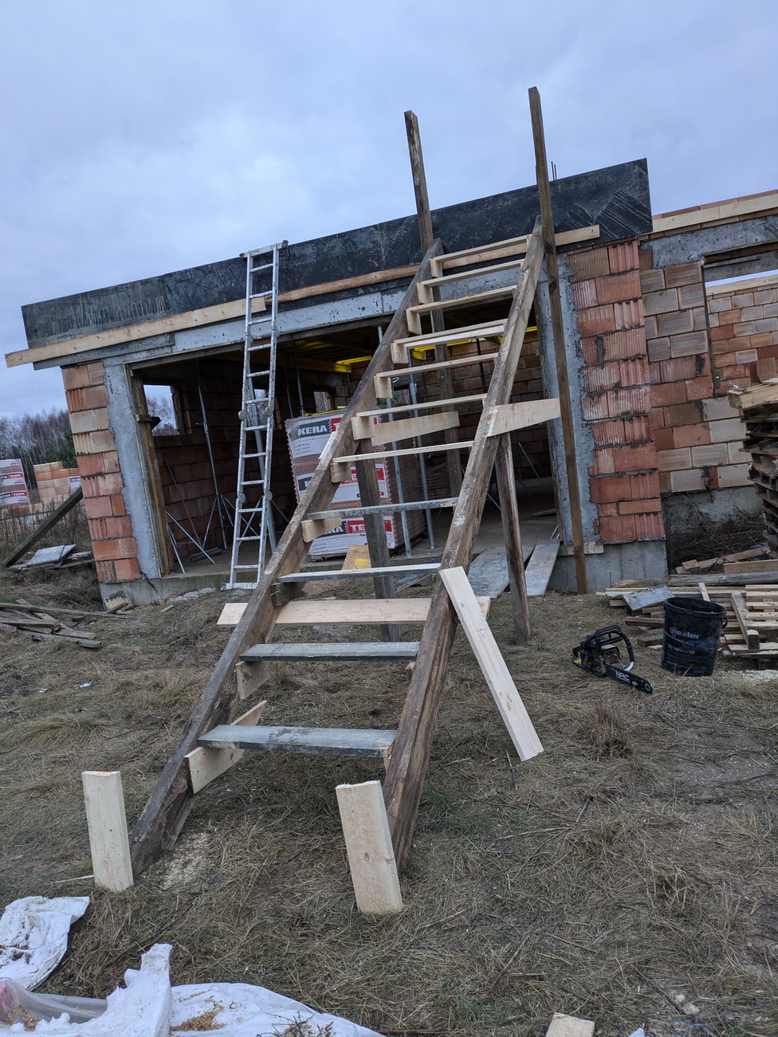 Surowy stan budowy domu z czerwonej cegły z widocznymi schodami z desek, metalową drabiną i narzędziami budowlanymi na ziemi.
