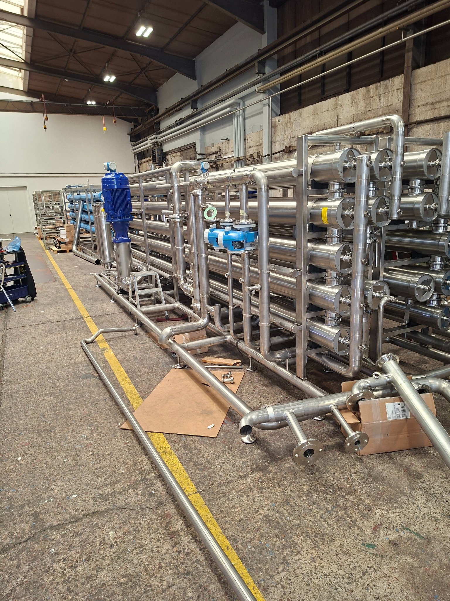 Kompleksowa instalacja przemysłowa z rur nierdzewnych, z widocznymi elementami łączeniowymi i silnikami, na tle hali produkcyjnej z żółtą linią na podłodze.