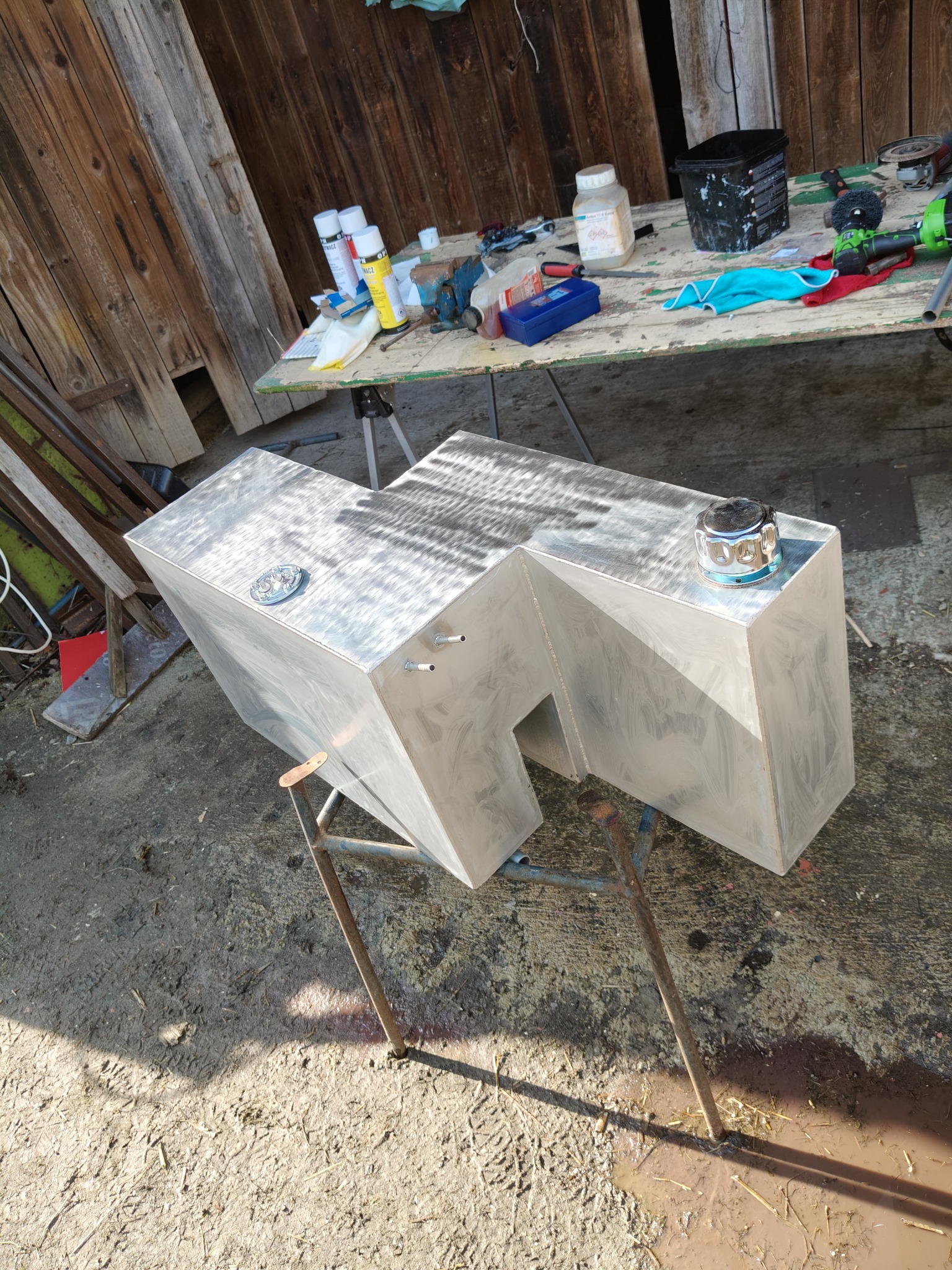 Dwa spawane, aluminiowe zbiorniki o nieregularnym kształcie, stojące na metalowych kozłach. W tle warsztatowy stół z narzędziami i drewniana ściana.