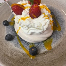 Restauracja Herbarium - Pojedyncza beza Pavlova zwieńczona bitą śmietaną, malinami, borówkami, listkiem mięty i sosem mango, podana na talerzu z szarą obwódką, nakładana łyżeczką.