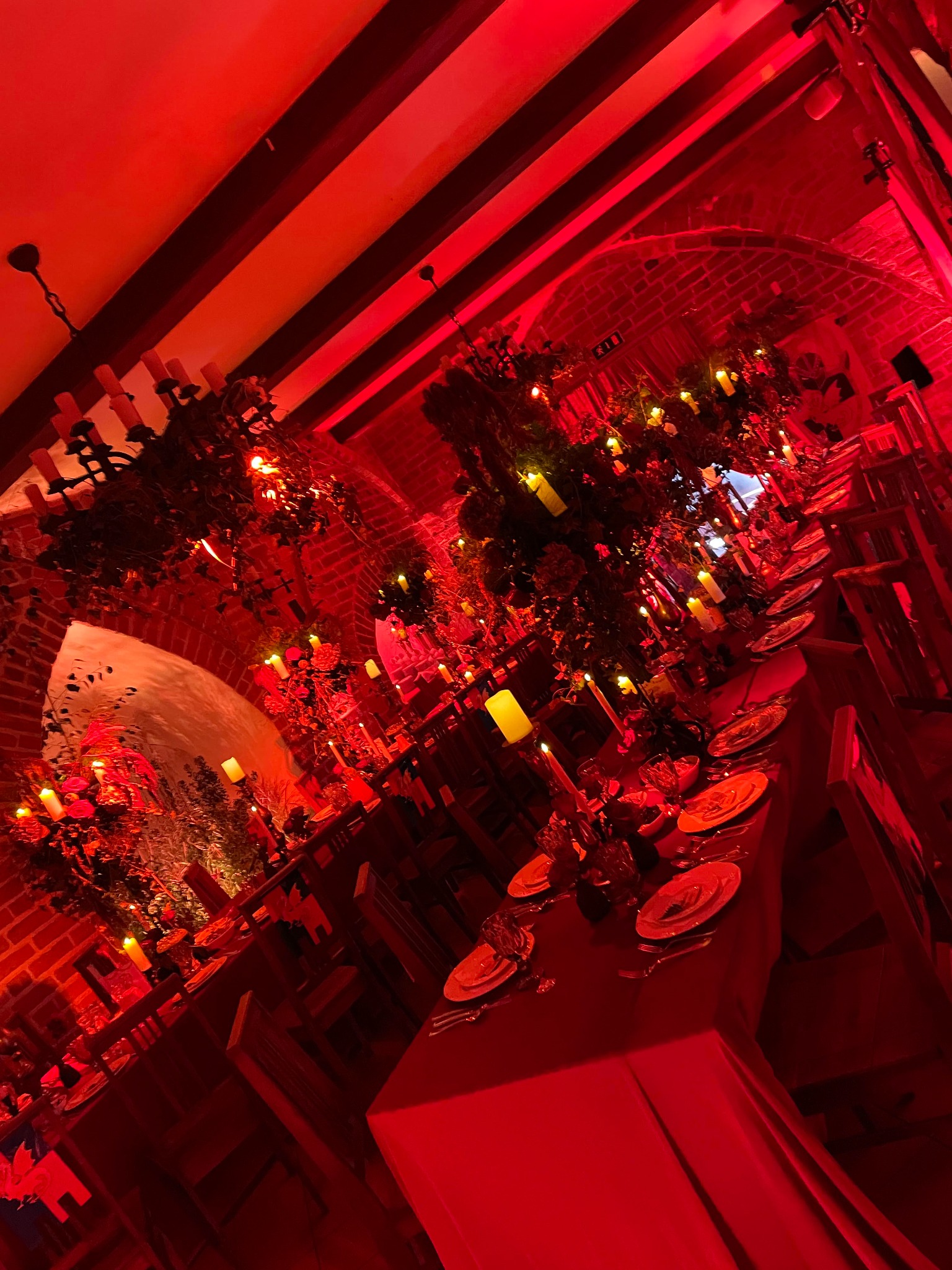 Uroczysta sala z długim stołem, nakrytym na czerwono, oświetlona czerwonym światłem, dekoracje kwiatowe i świece, ceglane ściany i łukowe przejścia, drewniane belki sufitowe, klimatyczne oświetlenie.