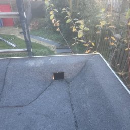 Kamil Podgórski - Dach pokryty papą z widocznym kominem i pęknięciami. W tle drabina, zieleń i budynki. Ujęcie z góry, lekko pochmurna pogoda.