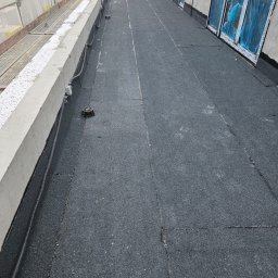 Kamil Podgórski - Dekarz w pracy na płaskim dachu pokrytym papą. Widoczne okna zabezpieczone folią, kable i elementy instalacji na dachu. Praca w toku.