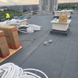 Kamil Podgórski - Prace na dachu płaskim z papy termozgrzewalnej: wentylacja, kable, narzędzia. W tle budynki mieszkalne i pochmurne niebo.