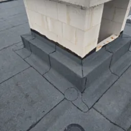 Szczelne krycie dachu papą wokół komina z cegły, z widocznymi detalami obróbki blacharskiej i odpływem wody. Na kominie leżą żółta kamizelka i biały kask.