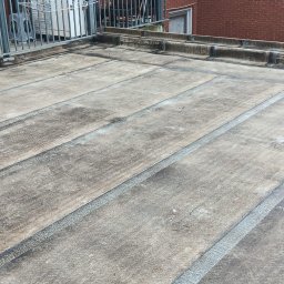 Kamil Podgórski - Płaski dach pokryty papą z widocznymi łączeniami, na tle balkonu z metalową barierką i fragmentem budynku z czerwonej cegły. Widoczne ślady użytkowania i zabrudzenia.