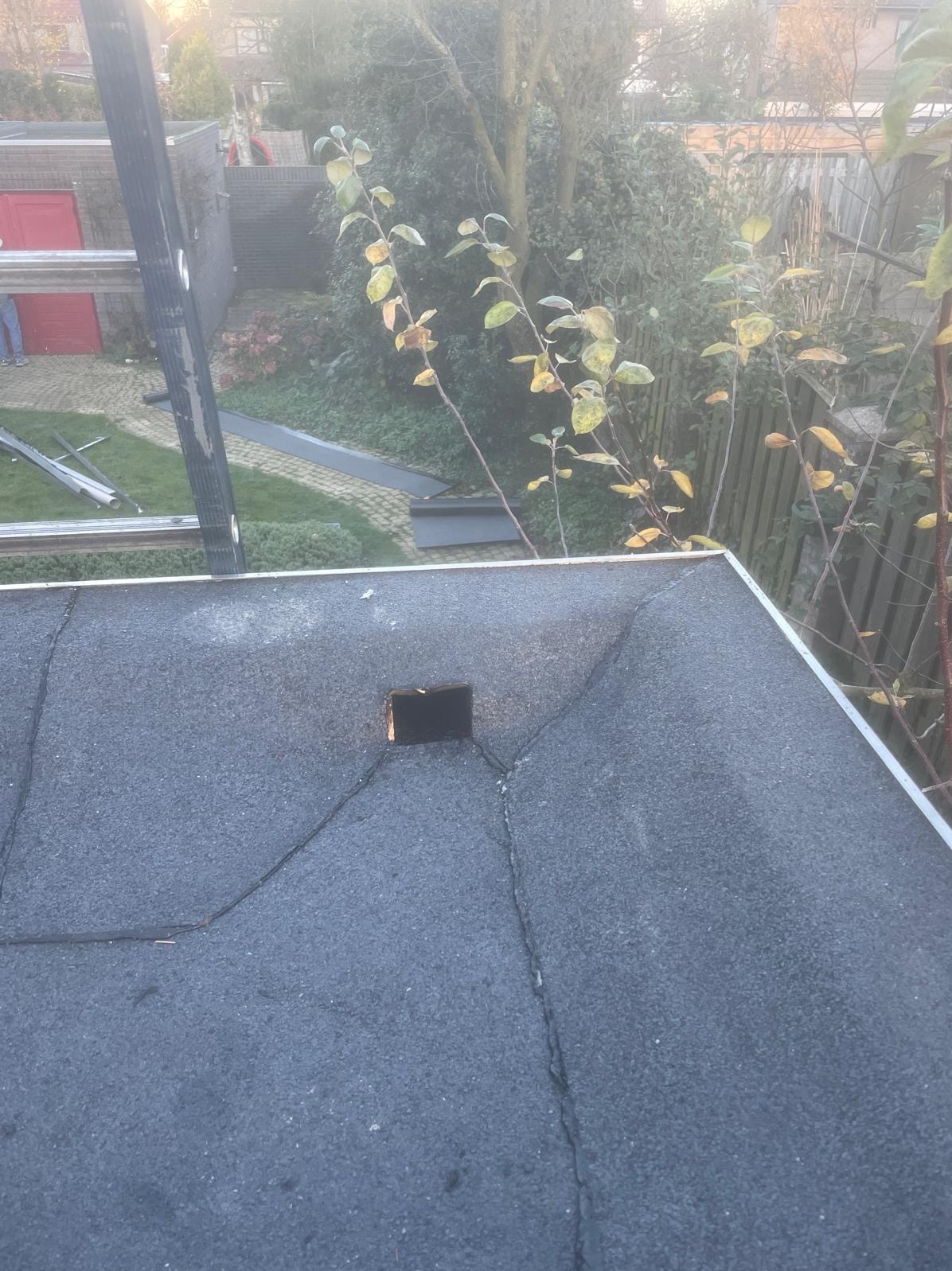 Dach pokryty papą z widocznym kominem i pęknięciami. W tle drabina, zieleń i budynki. Ujęcie z góry, lekko pochmurna pogoda.