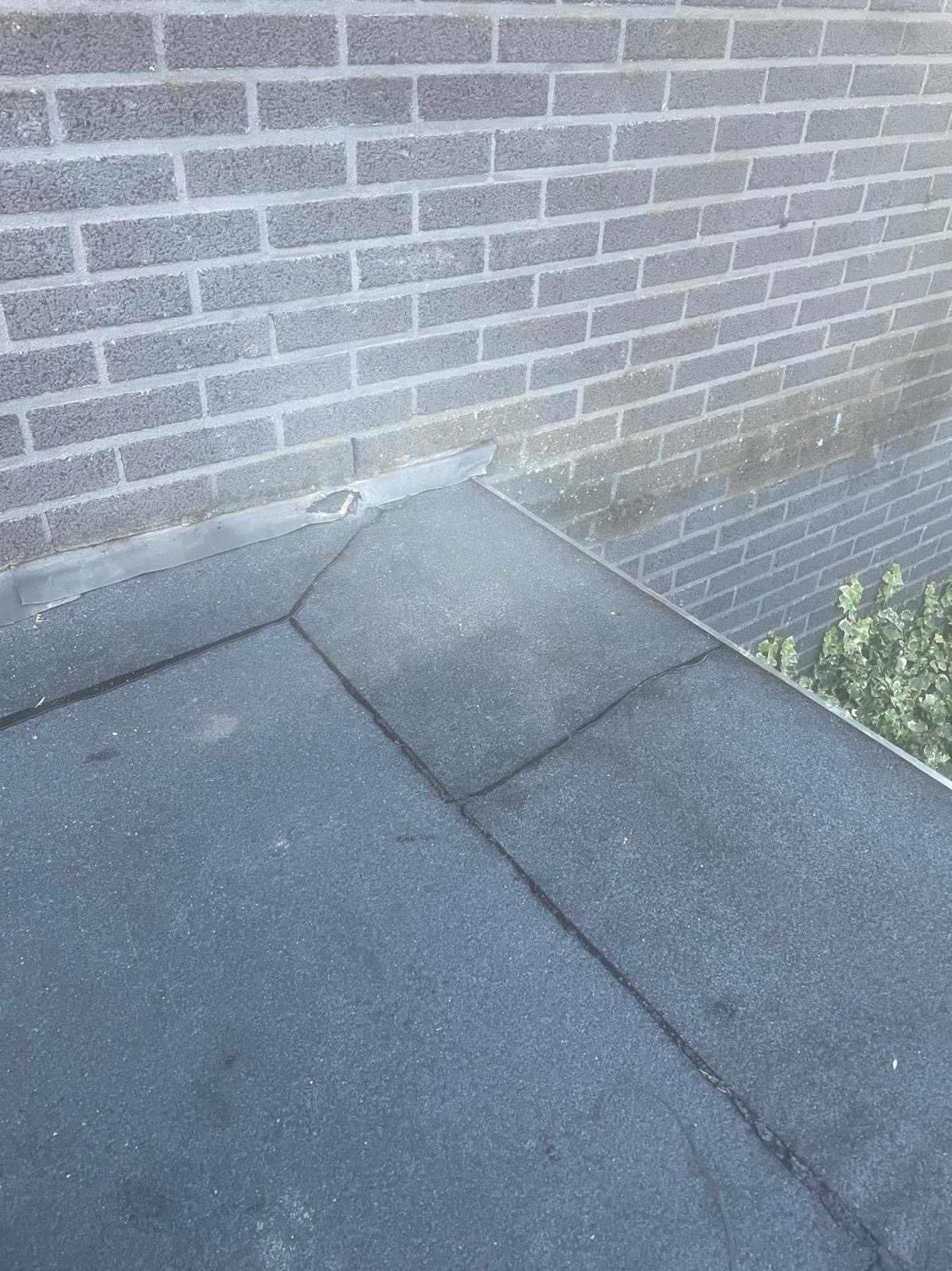 Płaski dach pokryty papą, przylegający do szarej, ceglanej ściany. Widoczne łączenia papy i fragment zieleni w tle. Ujęcie z góry.