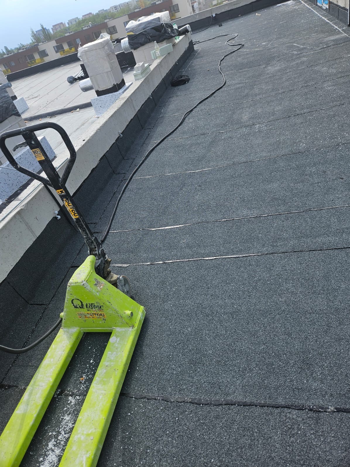 Gotowy dach pokryty papą termozgrzewalną z wózkiem paletowym. Widoczne zgrzewy papy i elementy konstrukcyjne dachu. Prace dekarskie w toku.