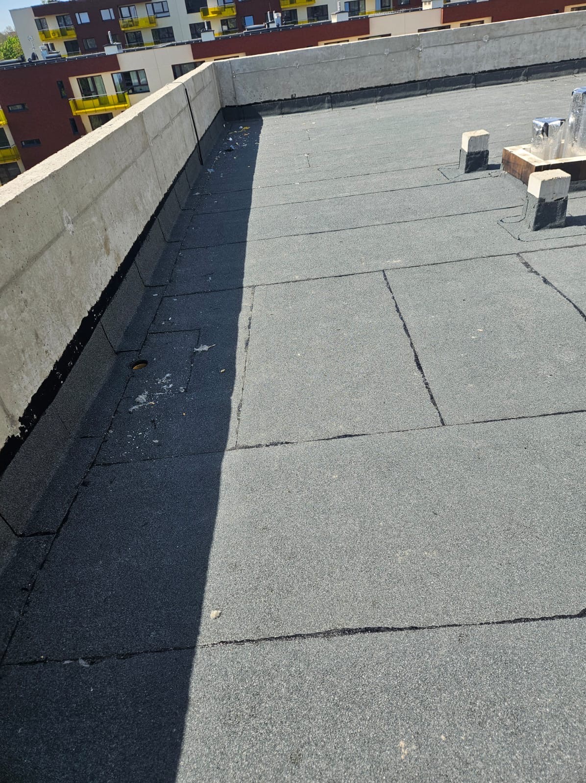 Płaski dach pokryty papą termozgrzewalną, z widocznymi łączeniami i elementami wentylacji. Betonowa attyka w tle, budynek mieszkalny w oddali.