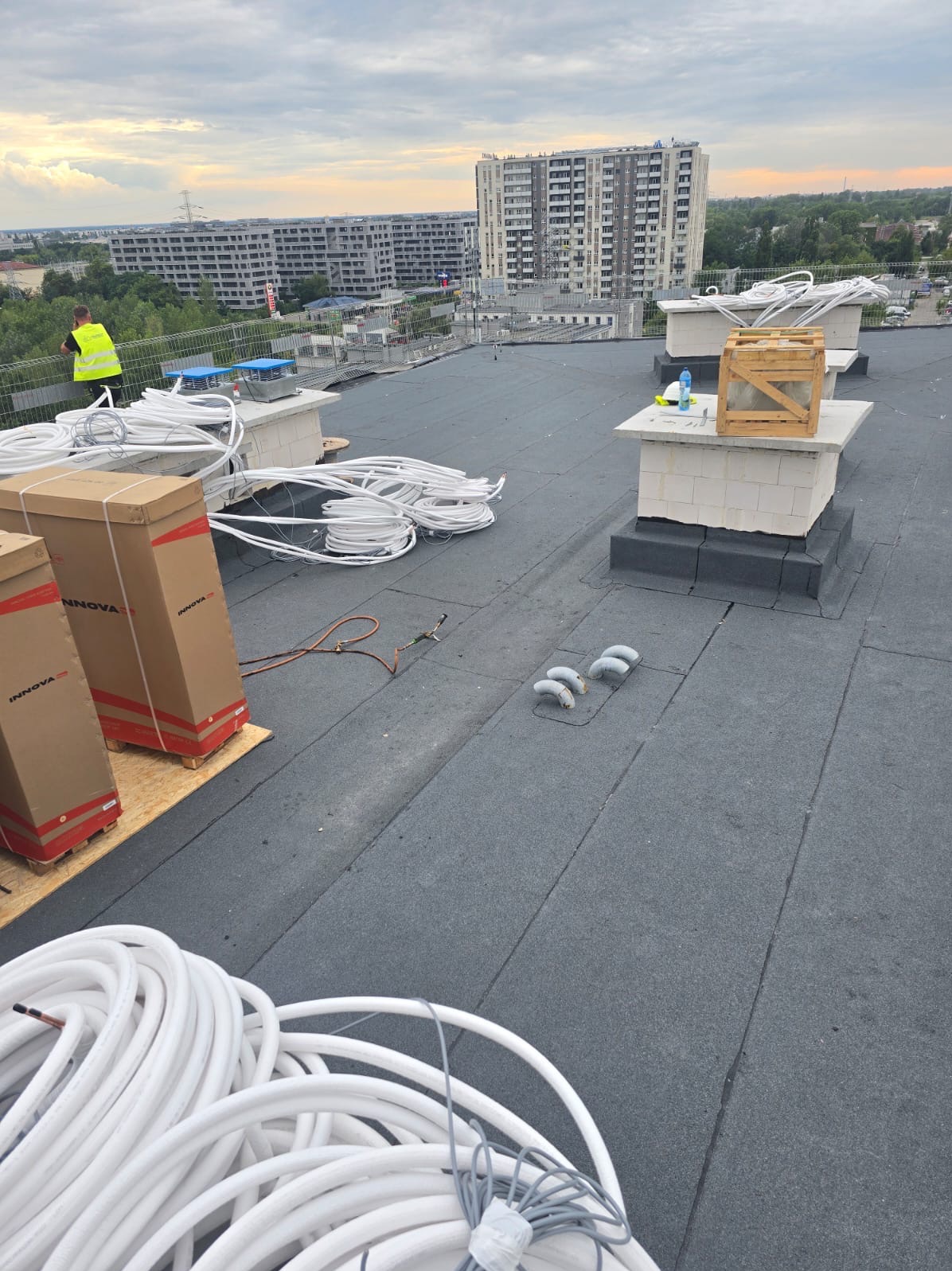 Prace na dachu płaskim z papy termozgrzewalnej: wentylacja, kable, narzędzia. W tle budynki mieszkalne i pochmurne niebo.