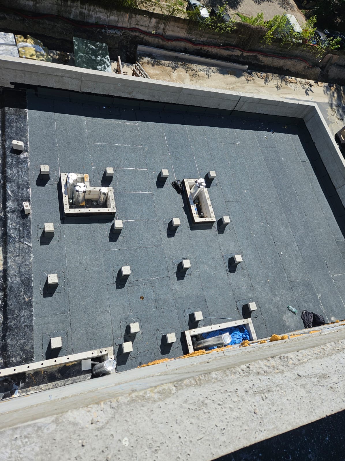 Nowe, szare pokrycie dachu płaskiego z betonowymi elementami i otworami wentylacyjnymi, widok z góry, w słoneczny dzień.