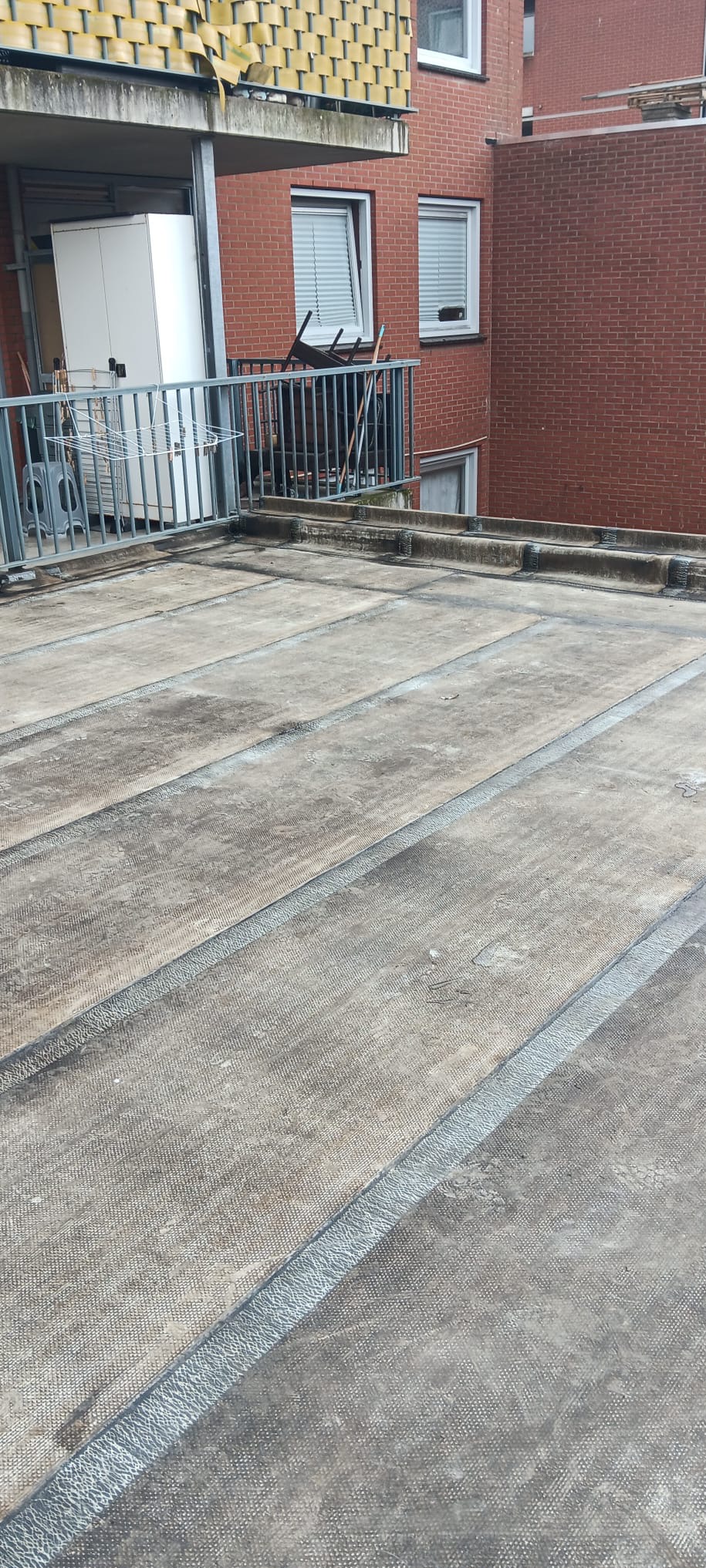 Płaski dach pokryty papą z widocznymi łączeniami, na tle balkonu z metalową barierką i fragmentem budynku z czerwonej cegły. Widoczne ślady użytkowania i zabrudzenia.