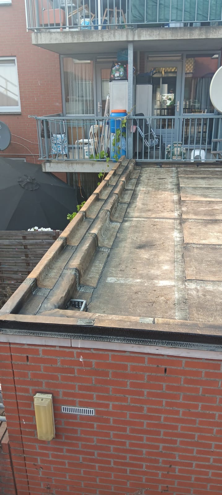 Płaski dach pokryty papą termozgrzewalną z obróbkami blacharskimi, widok z góry na tle ceglanego budynku z balkonem i anteną satelitarną.