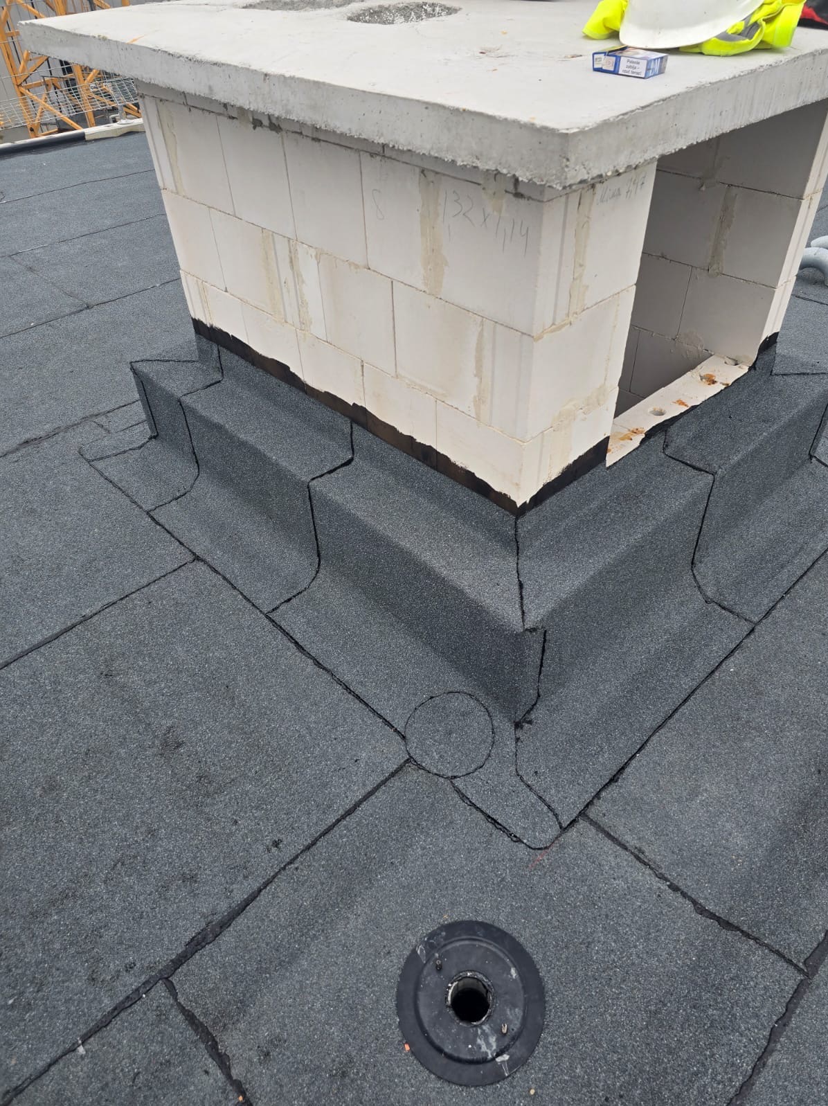 Szczegółowe krycie dachu papą wokół komina w Leszczawie Dolnej. Widoczny system odprowadzania wody. Biały kask i odblaskowa kamizelka leżą na kominie.