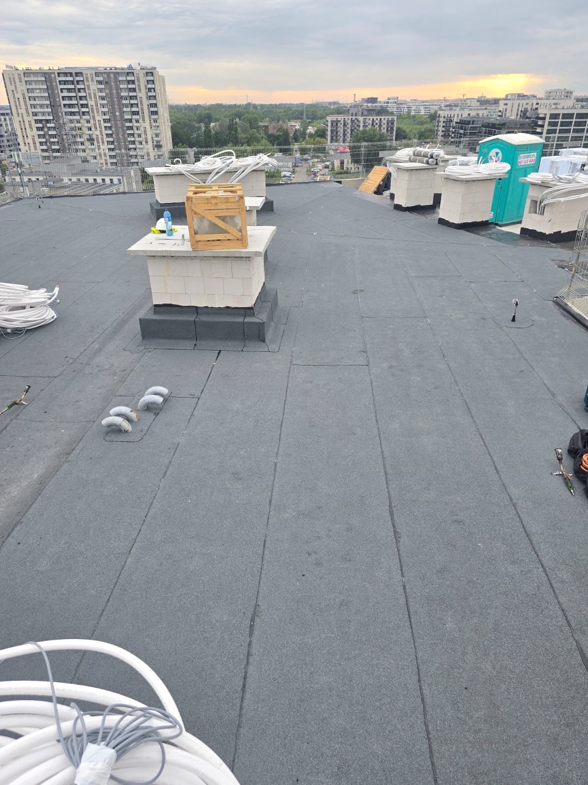Wykończony płaski dach pokryty papą termozgrzewalną z kominami wentylacyjnymi, w tle budynki mieszkalne i zachodzące słońce. Widoczne akcesoria dekarskie.