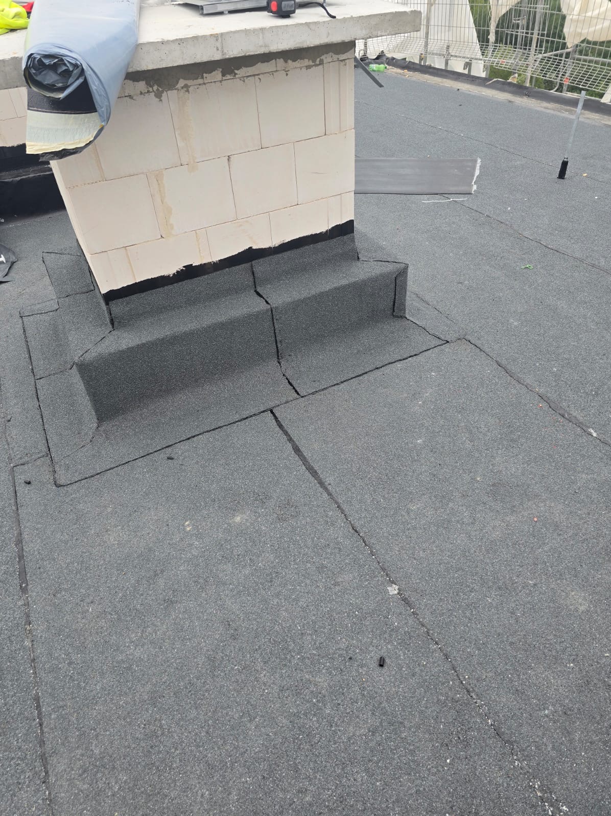 Szczelne krycie dachu papą wokół komina z cegieł. Detal wykonania obróbki komina na dachu płaskim. Nowoczesna hydroizolacja zgrzewana.