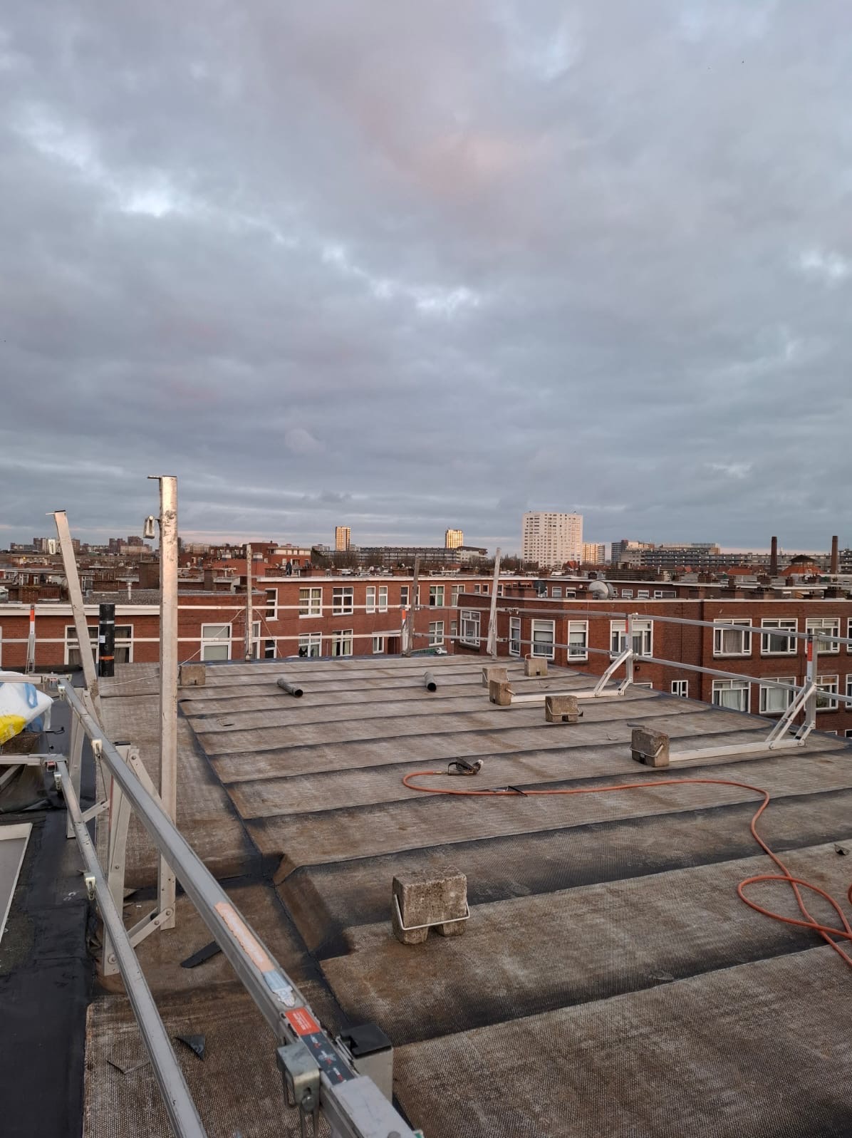 Płaski dach pokryty papą w trakcie prac, widoczne narzędzia, elementy rusztowania oraz budynki w tle. Kąt ujęcia z góry.