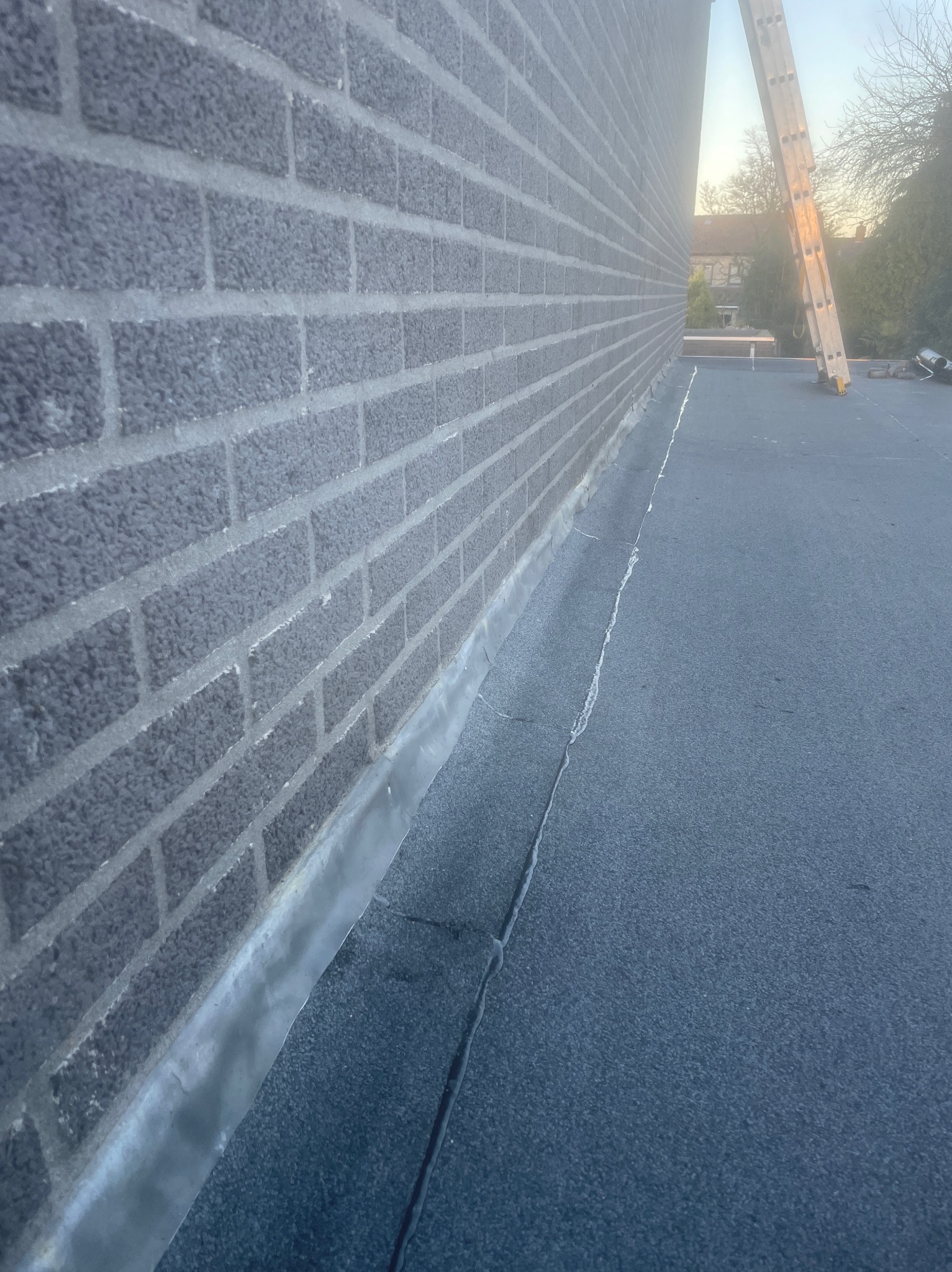 Szczegółowe ujęcie płaskiego dachu pokrytego papą, z widoczną ścianą z szarej cegły i fragmentem drabiny opartą o dach. Widoczne łączenia papy.