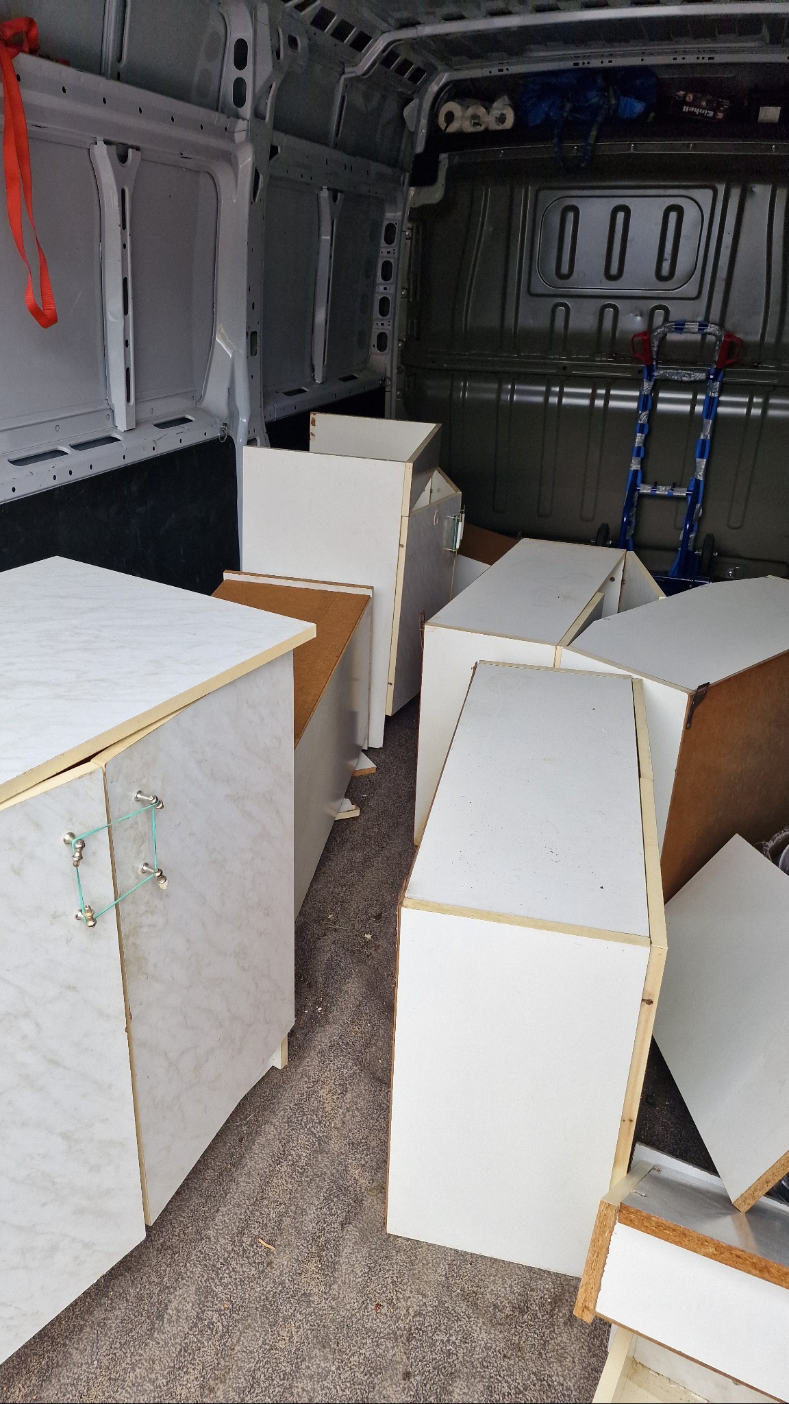 Wnętrze furgonetki wypełnionej białymi szafkami z marmurkowym wzorem, przygotowanymi do transportu. W tle widoczny wózek transportowy i zabezpieczenia ładunku.