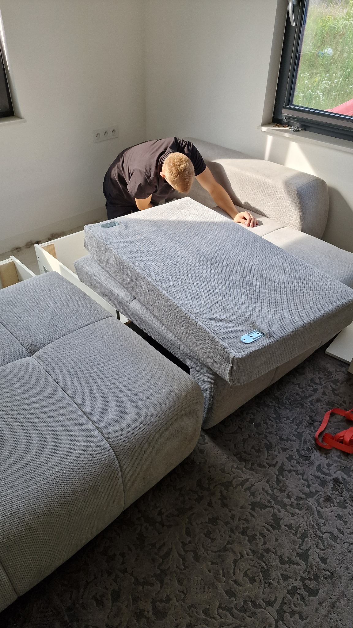 Mężczyzna montuje szarą sofę modułową w jasnym pokoju z oknem; widoczne elementy konstrukcyjne i poduszki sofy na wzorzystym dywanie.