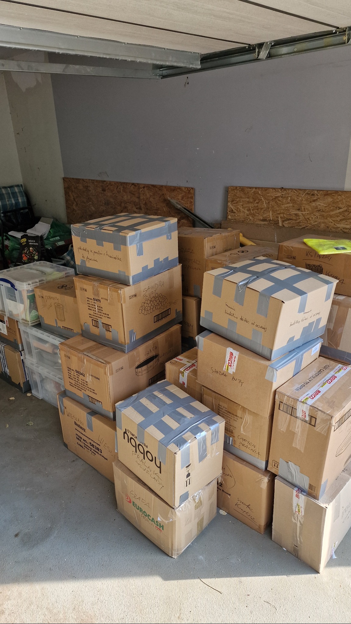 Stos kartonowych pudeł z taśmą, niektóre z odręcznymi napisami, zgromadzone w garażu, czekające na transport. Widoczny bałagan i przygotowania do przeprowadzki.