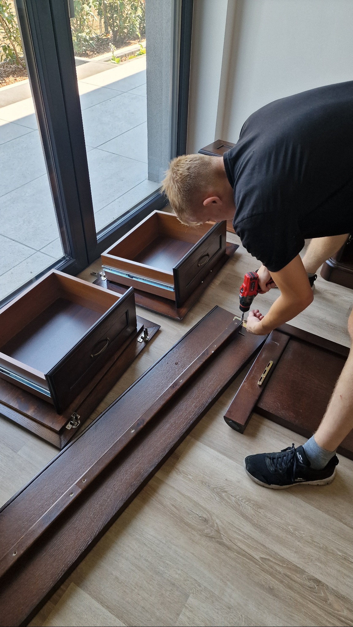 Mężczyzna skręca meble w pomieszczeniu z dużym oknem, używając wkrętarki. Widoczne elementy szuflad i boku szafy, podłoga imitująca drewno.
