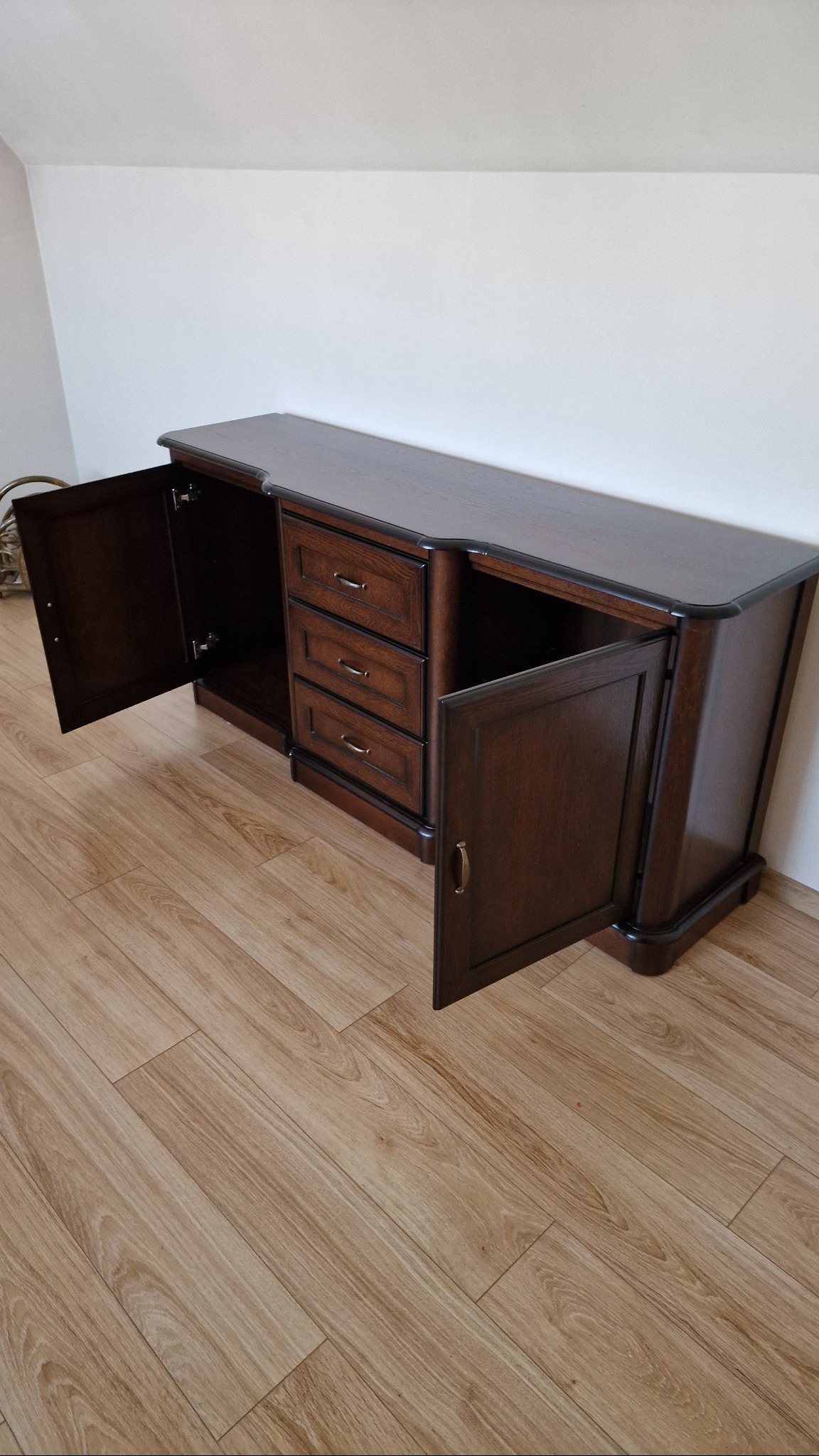 Drewniana komoda z trzema szufladami i otwieranymi drzwiczkami, ustawiona na podłodze z paneli, widoczna na tle białej ściany.