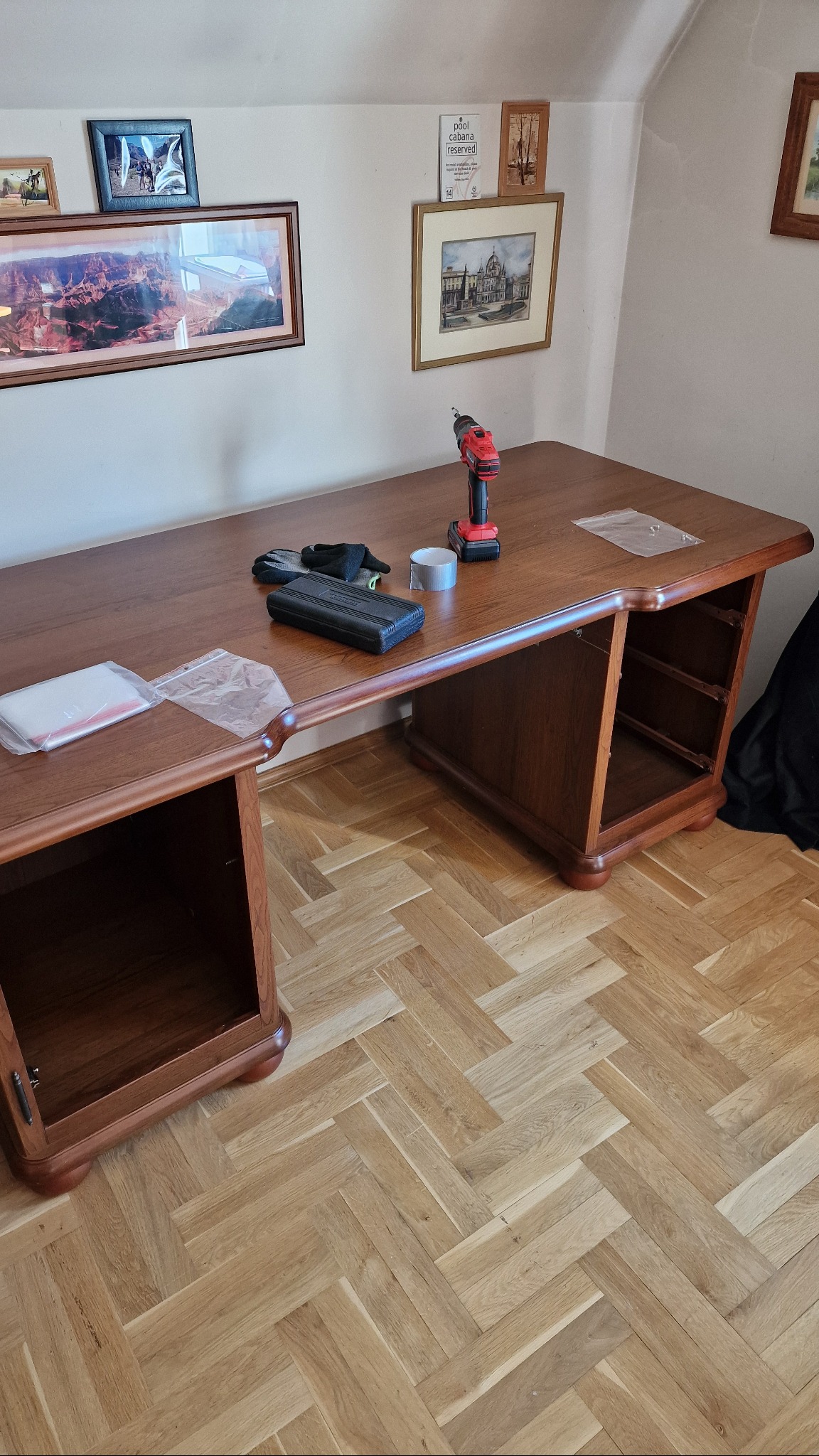 Demontaż biurka z drewnianym blatem i nogami. Na blacie wiertarka, taśma, rękawice i pudełko. Parkiet w jodełkę. Przygotowanie do transportu.