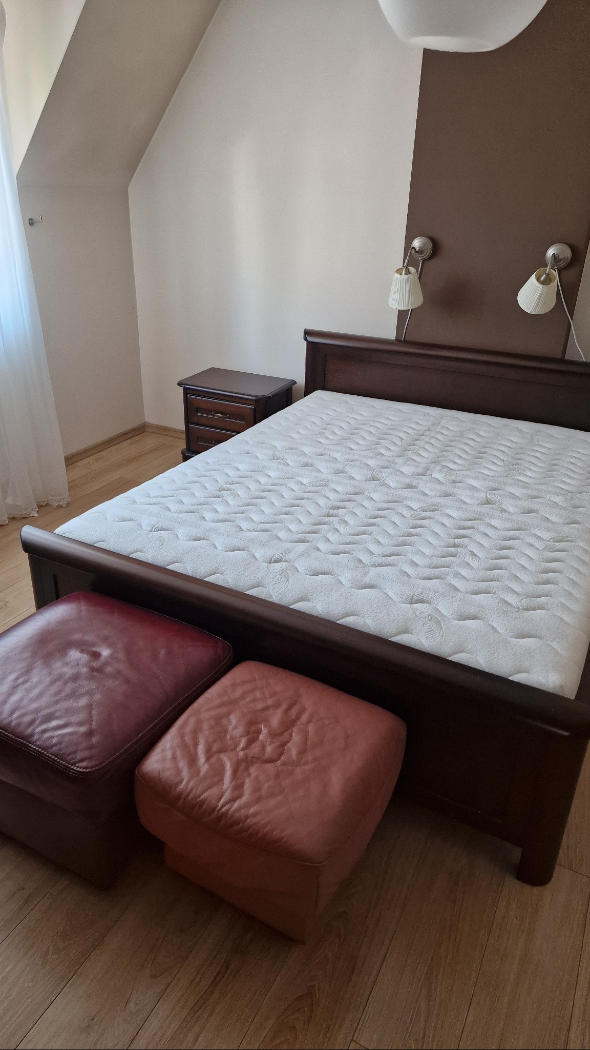 Sypialnia z drewnianym łóżkiem z materacem i dwoma pufami w odcieniach czerwieni. Na ścianie lampki nocne.