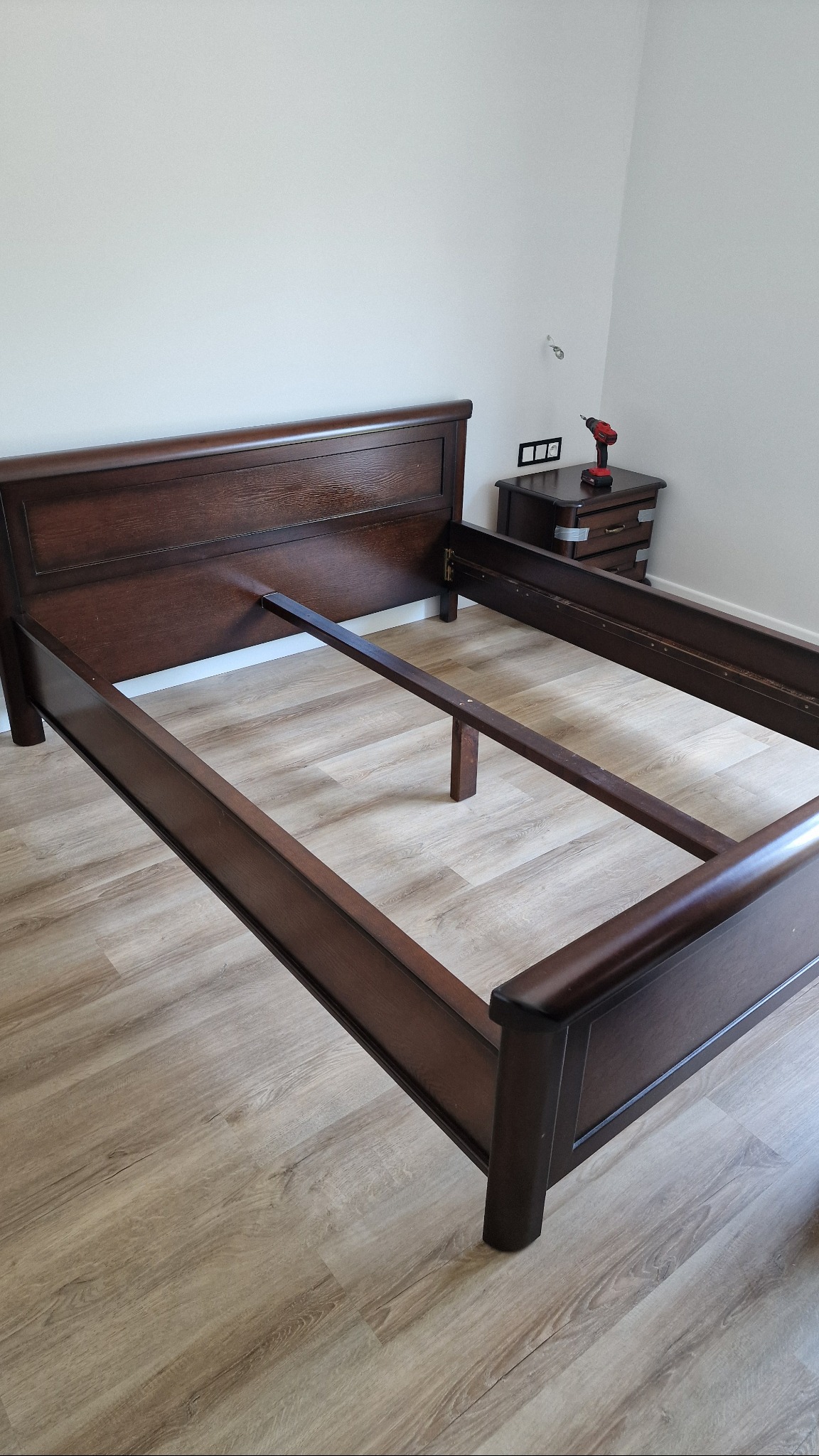 Demontaż łóżka z ciemnego drewna, z widoczną wkrętarką na szafce nocnej. Etap przygotowania do transportu, jasna podłoga i ściany.