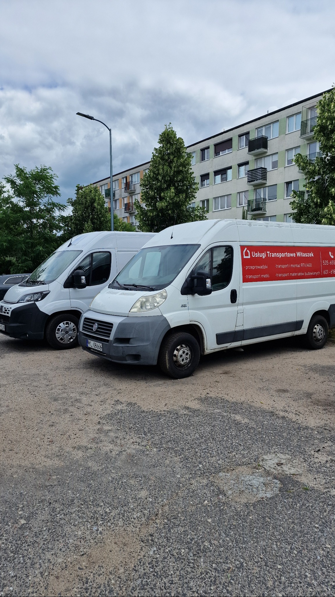 Dwa białe busy firmy transportowej Witaszek zaparkowane na szutrowym parkingu, na tle bloku mieszkalnego i drzew. Na busach widoczne logo i zakres usług.