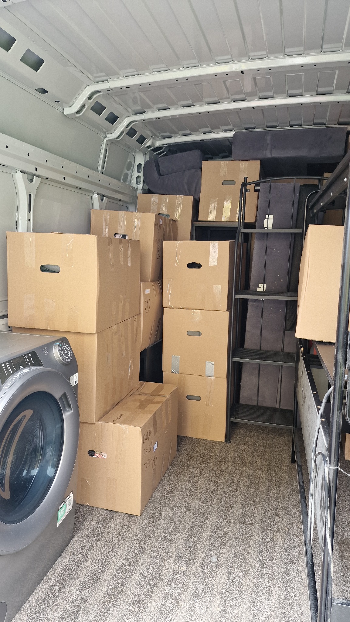 Wnętrze furgonetki załadowane kartonami, pralką i meblami w trakcie relokacji. Widoczne zabezpieczone przedmioty gotowe do transportu.