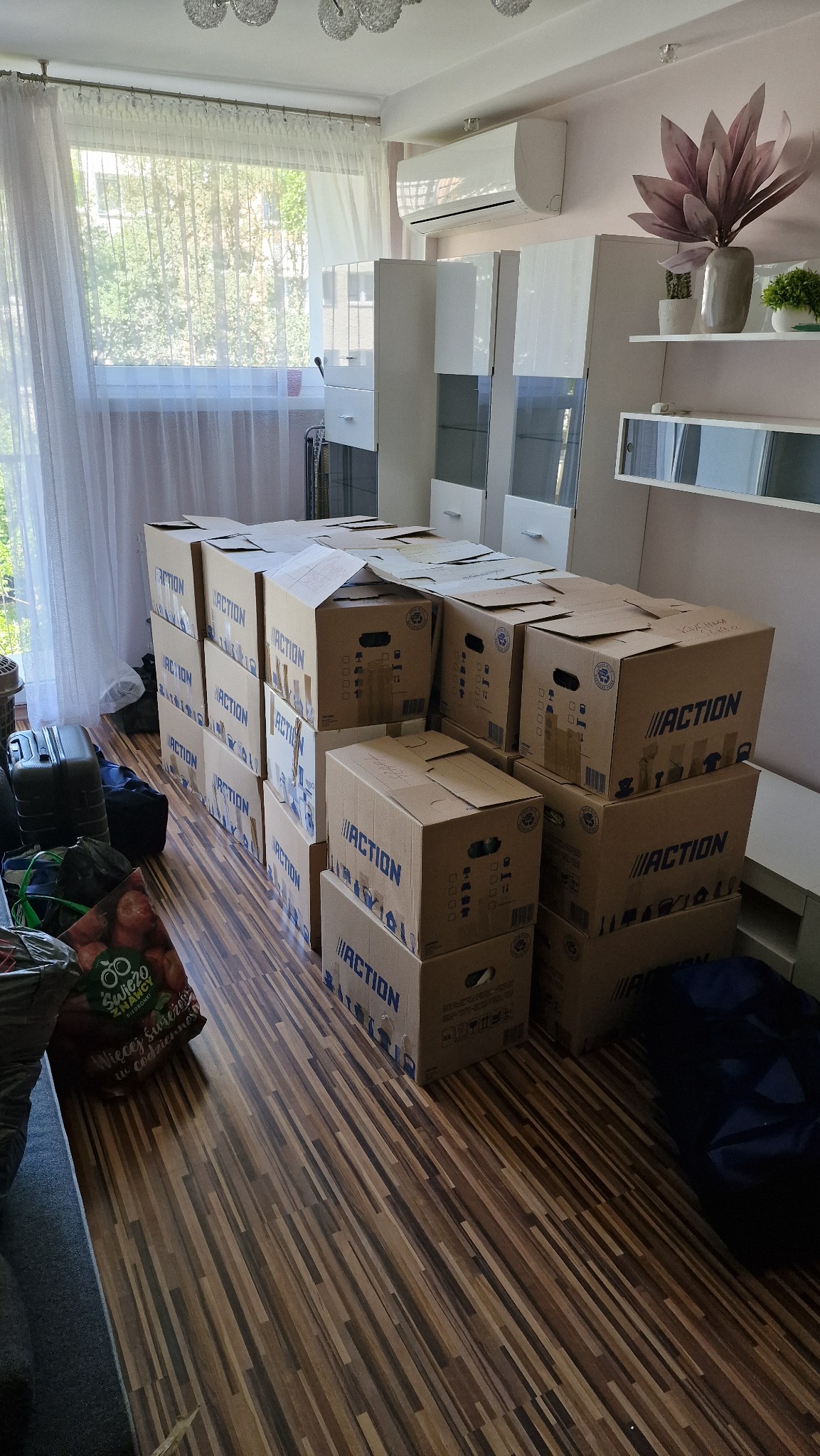 Pokój z kartonami marki Action ułożonymi w stosy, przygotowanymi do transportu. Widoczne torby i walizki, sugerujące pakowanie dobytku.