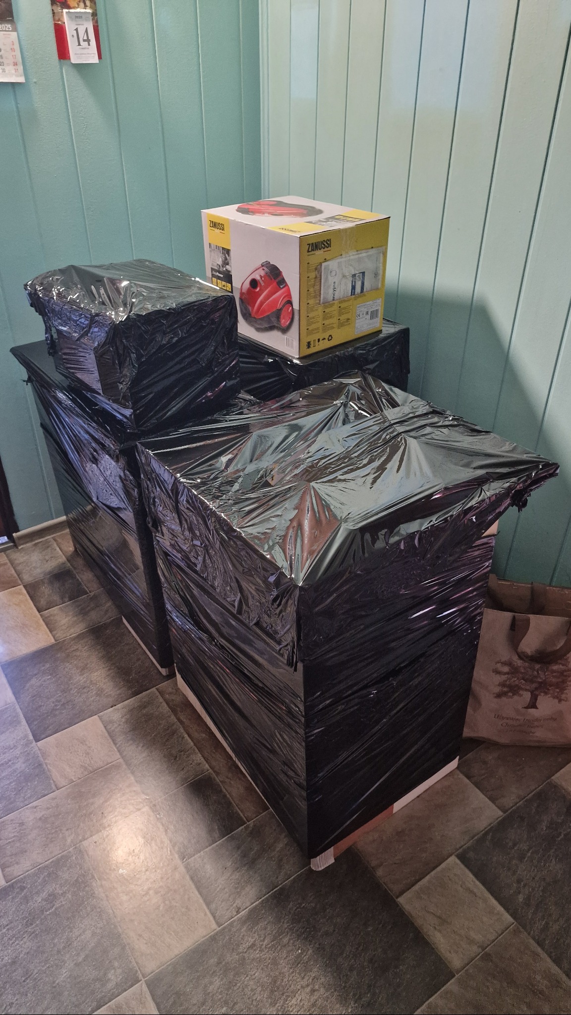 Zabezpieczone folią meble i karton z odkurzaczem Zanussi, gotowe do transportu. Ułożone na podłodze z płytek, przy zielonej ścianie.