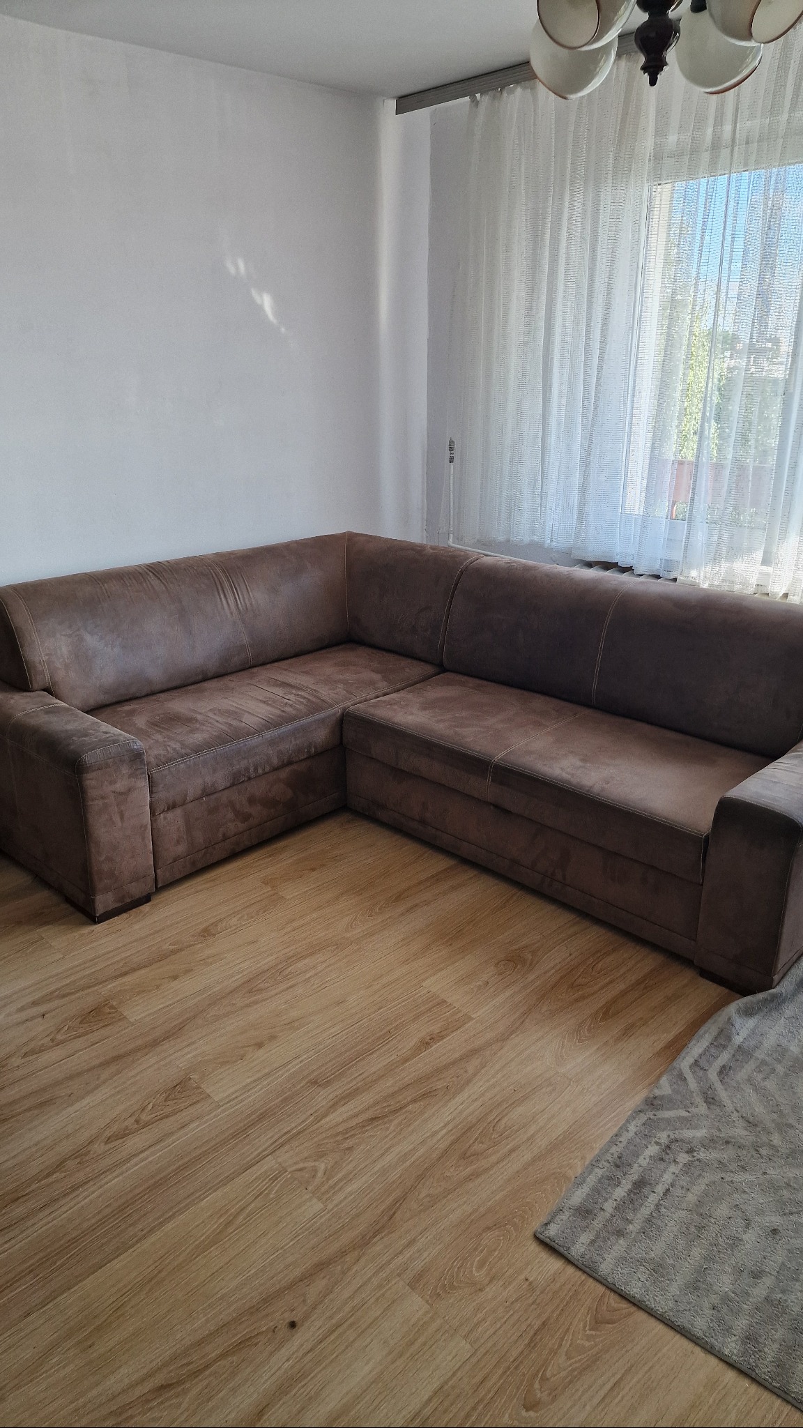 Brązowa, narożna sofa z mikrofibry w jasnym pokoju z drewnianą podłogą i zasłoniętym oknem. Częściowo widoczny dywan. Widok na wnętrze po przeprowadzce.