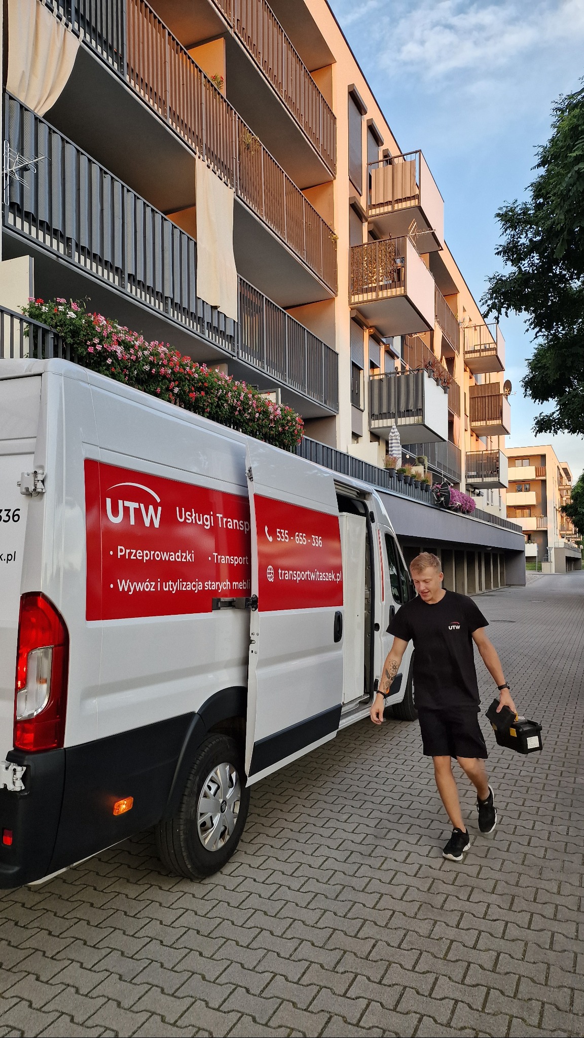 Biały van z otwartymi drzwiami i logo firmy transportowej UTW na tle nowoczesnego budynku mieszkalnego. Mężczyzna w czarnym ubraniu niesie skrzynkę narzędziową.