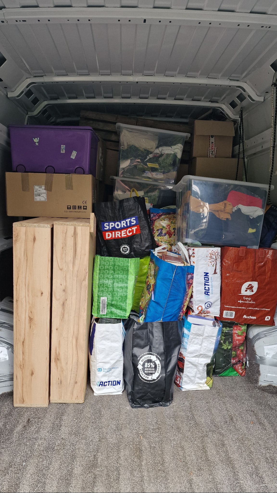 Wnętrze busa załadowane kartonami, pudłami, torbami z zakupami i dwoma drewnianymi elementami, przygotowane do transportu. Widok z tyłu pojazdu.