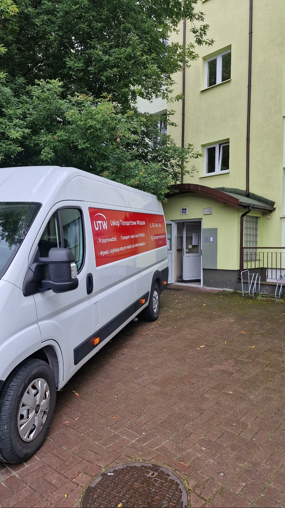 Biały bus firmy transportowej UTW na brukowanym podjeździe przed budynkiem z numerem 40A. Na busie czerwone logo i dane kontaktowe. Drzewo w tle.