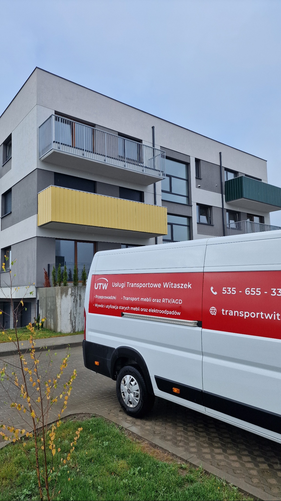 Biała furgonetka z czerwonym logo firmy przeprowadzkowej 'UTW Usługi Transportowe Witaszek' na tle nowoczesnego bloku mieszkalnego.