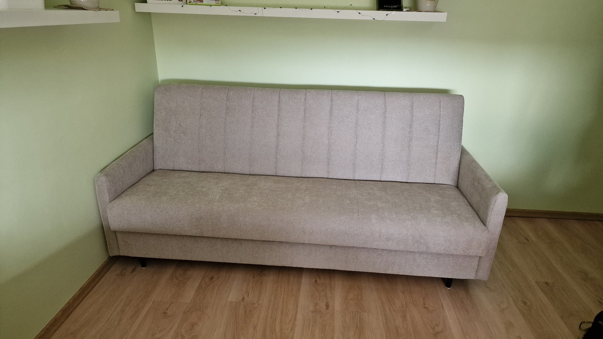 Jasnoszara sofa z pionowymi przeszyciami na tle pistacjowej ściany i drewnianej podłogi. Minimalistyczny design i prosta forma sofy.