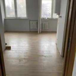 Jasne panele podłogowe imitujące drewno w pustym pokoju, widoczne dwa okna z grzejnikami i fragment bojlera gazowego, ściany szare, widoczne ślady po pracach wykończeniowych.