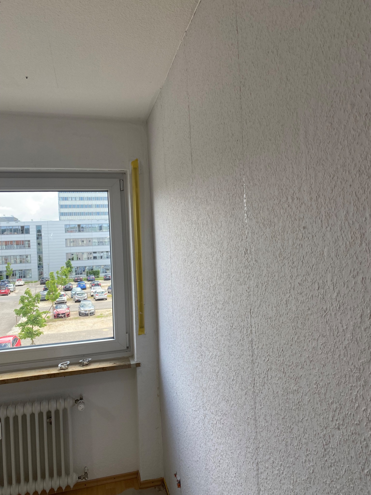 Pokój w trakcie remontu: ściana z nierówną fakturą przygotowana do szpachlowania, okno z widokiem na parking, grzejnik pod oknem.