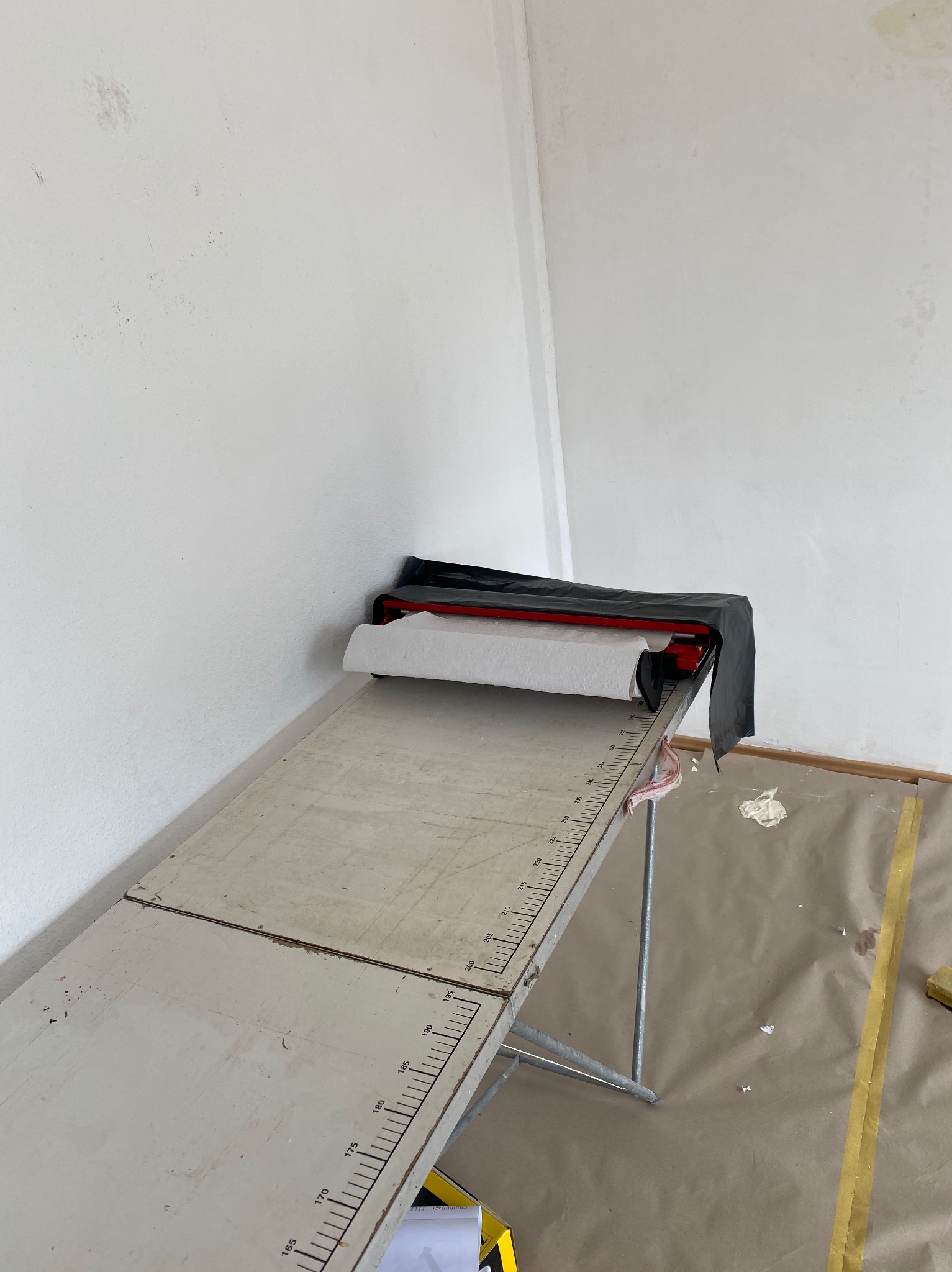 Przygotowania do montażu sufitu napinanego: rolka folii na stole z miarką, zabezpieczona czarną osłoną, widoczne ślady zabrudzeń na ścianie i podłodze zabezpieczonej papierem malarskim.