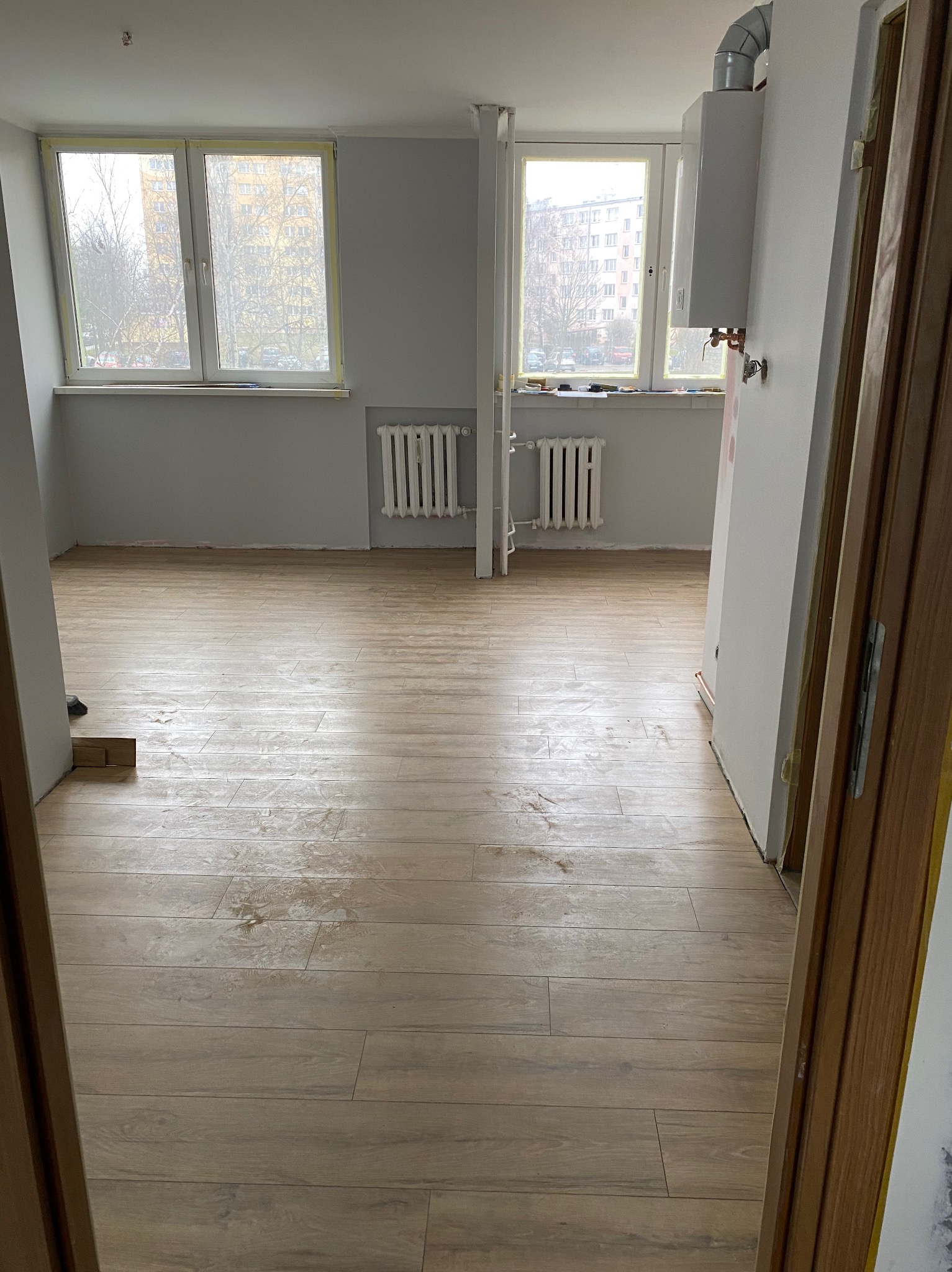 Jasne panele podłogowe imitujące drewno w pustym pokoju, widoczne dwa okna z grzejnikami i fragment bojlera gazowego, ściany szare, widoczne ślady po pracach wykończeniowych.