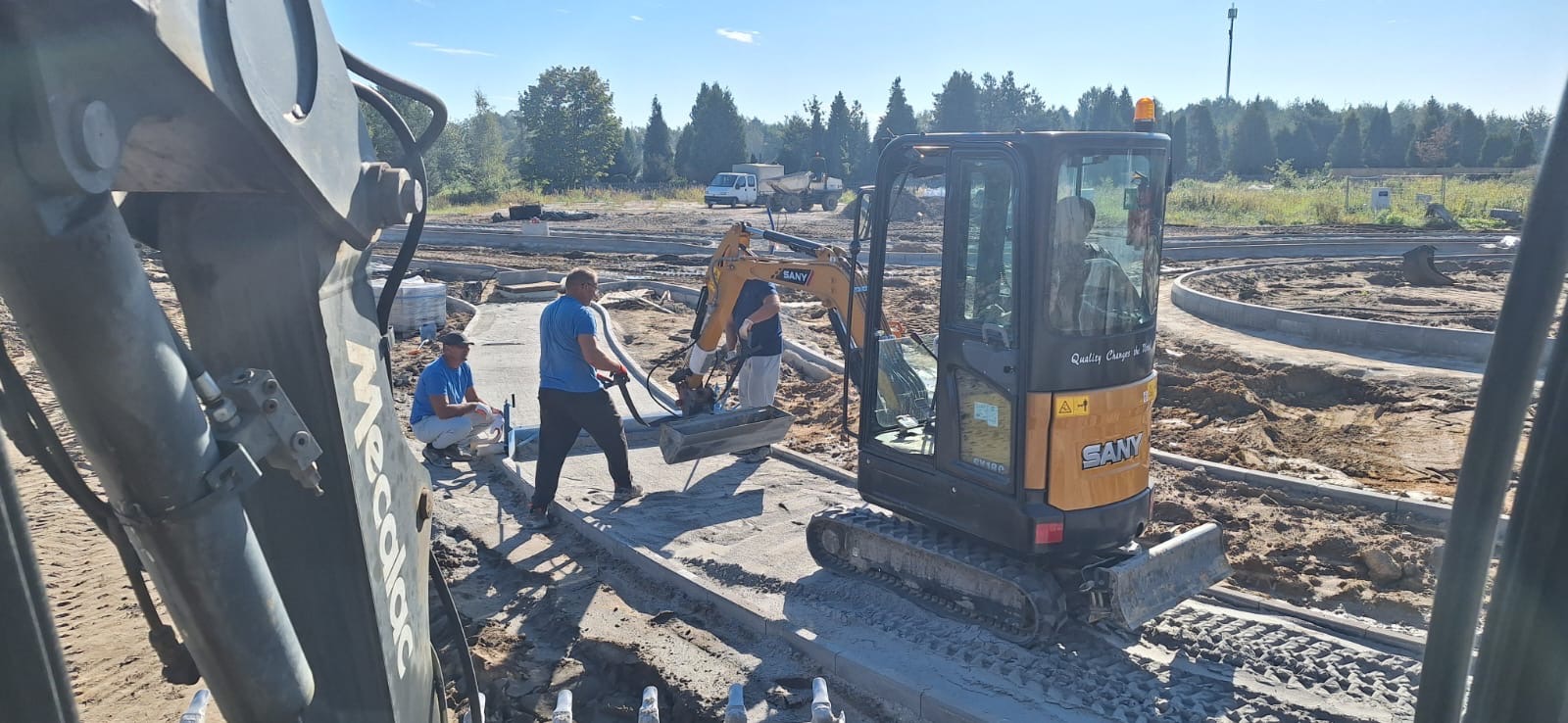 Prace ziemne przy użyciu minikoparki SANY na terenie budowy, widoczni robotnicy, przygotowanie podłoża pod chodnik z kostki brukowej, w tle drzewa i błękitne niebo.