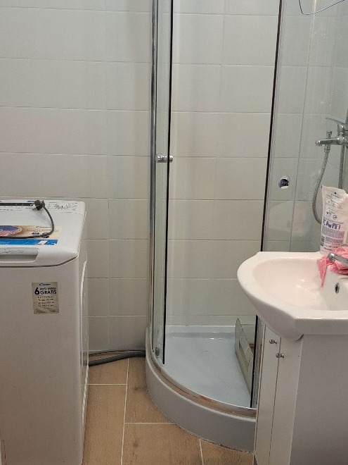 Umywalka z szafką, kabina prysznicowa z przezroczystymi drzwiami i pralka w małej łazience z jasnymi płytkami i brązową podłogą.