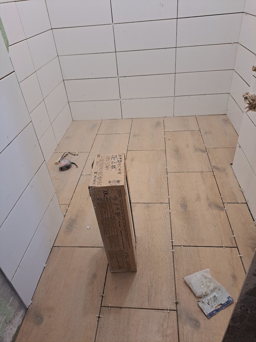 Wnętrze łazienki w trakcie prac wykończeniowych: ułożone płytki podłogowe imitujące drewno z krzyżykami dystansowymi, białe płytki ścienne, karton z płytkami i narzędzia na podłodze.