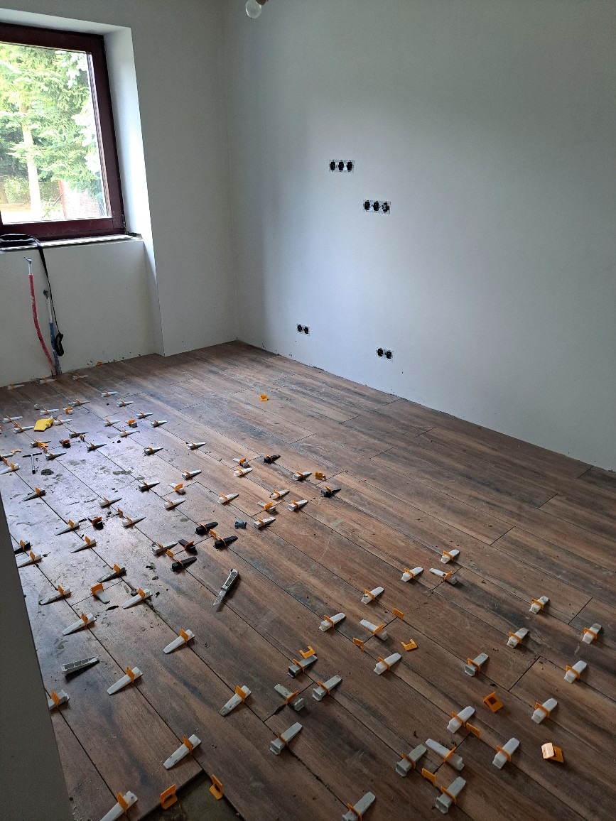 Układanie drewnopodobnych płytek podłogowych z użyciem klipsów poziomujących w jasnym pomieszczeniu z oknem i przygotowanymi gniazdkami elektrycznymi.